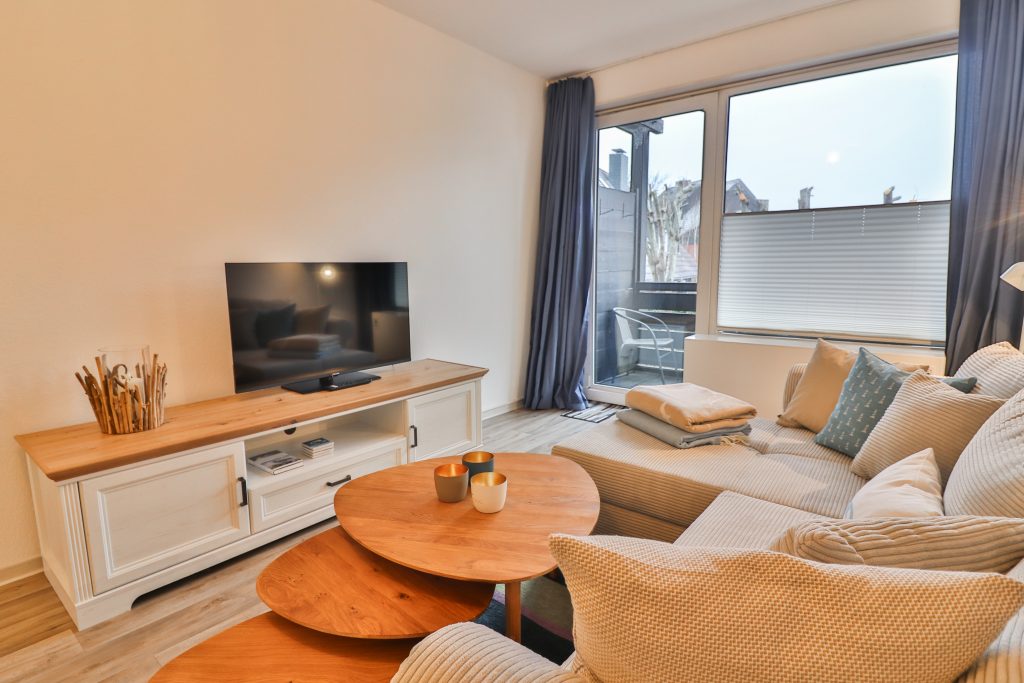 Düne 53 Grad Wohnzimmer mit beigen Cordsofa, Wohnzimmertischen TV Kommode und einem Fernseher darauf.