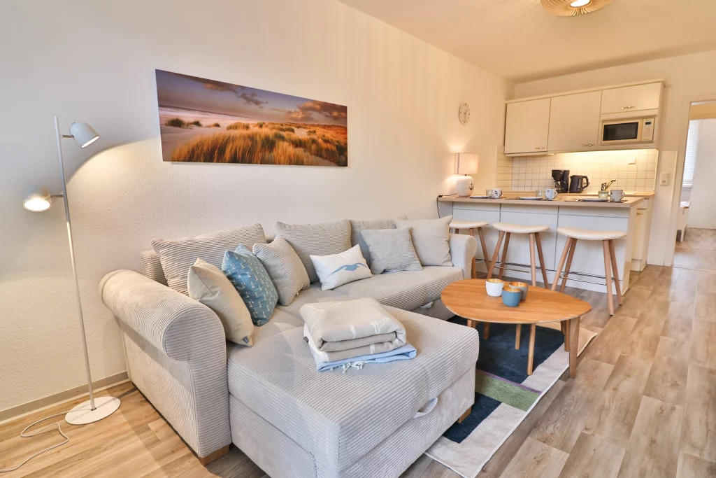 Düne 53 Grad Wohnzimmer mit beigen Cordsofa, Wohnzimmertischen und Blick auf die weiße Küche mit Sitzgelegenheiten.